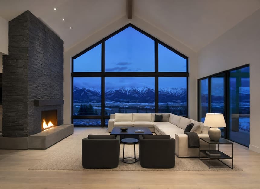 The finished interior of a new Montana home at dusk: vaulted great room, black steel windows, stone fireplace and mountains outside the window wall.