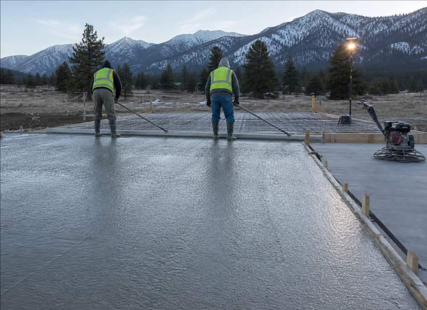 A concrete crew pulling a screed across a freshly poured house foundation slab on a Montana lot at first light.